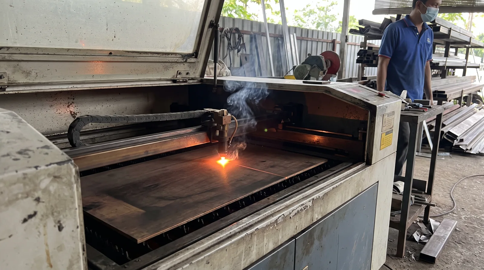 CO2 laser cutting machine engraving a flat sheet inside a workshop, bright beam spot and fine wisp of smoke