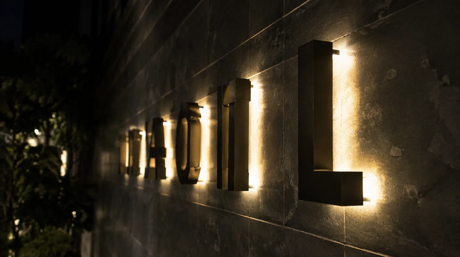 Back-lit stainless steel 3D letters on spacers with a warm halo glow on a dark stone wall