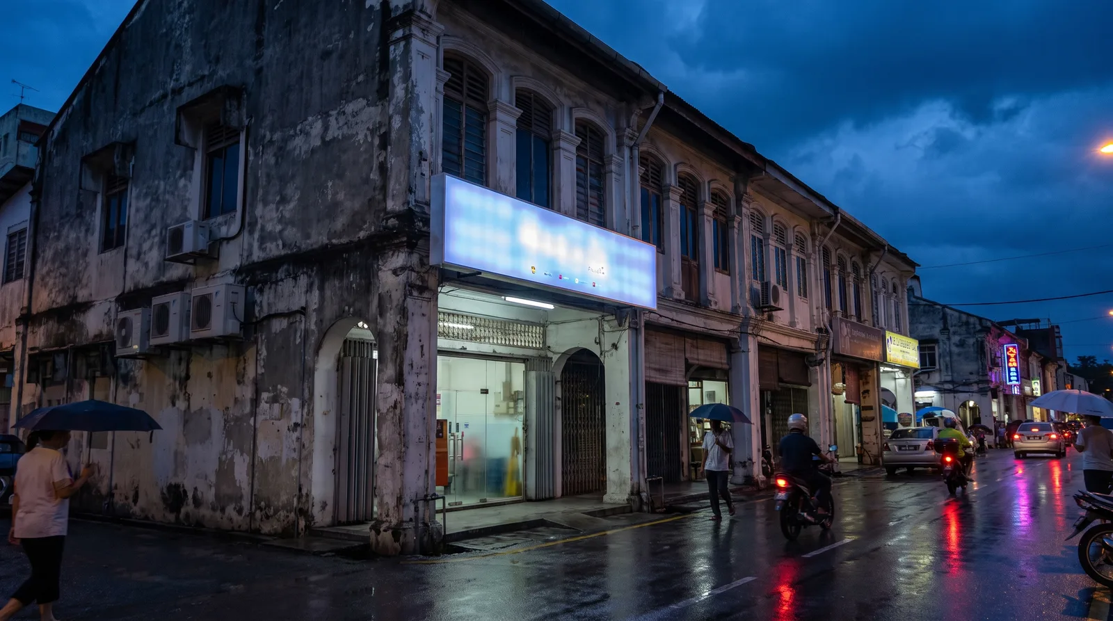 illuminated acrylic lightbox sign glowing above a Malaysian shoplot at dusk, blue hour sky and wet road reflections