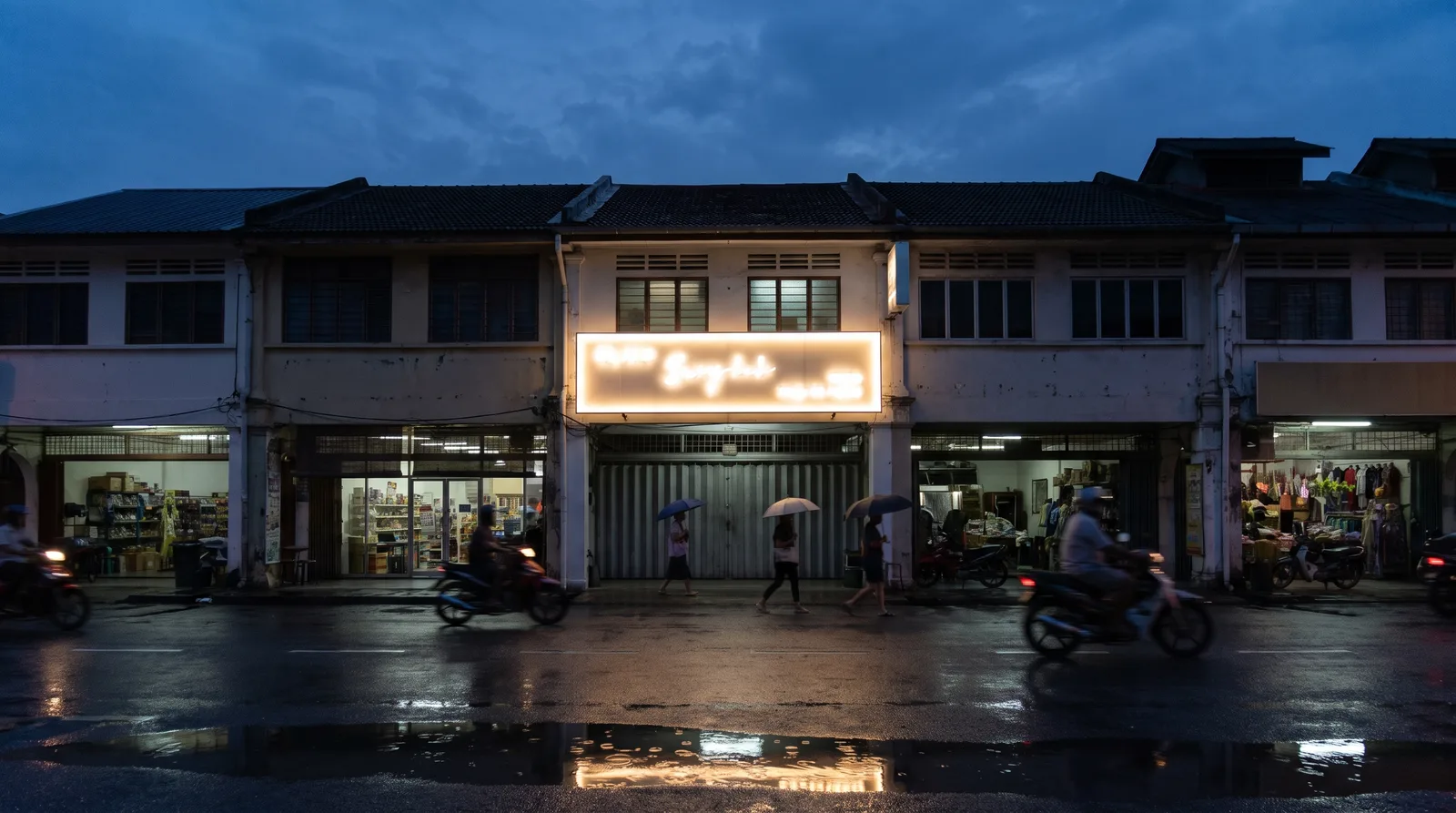 illuminated acrylic lightbox sign glowing above a Malaysian shoplot at dusk, blue hour sky and wet road reflections