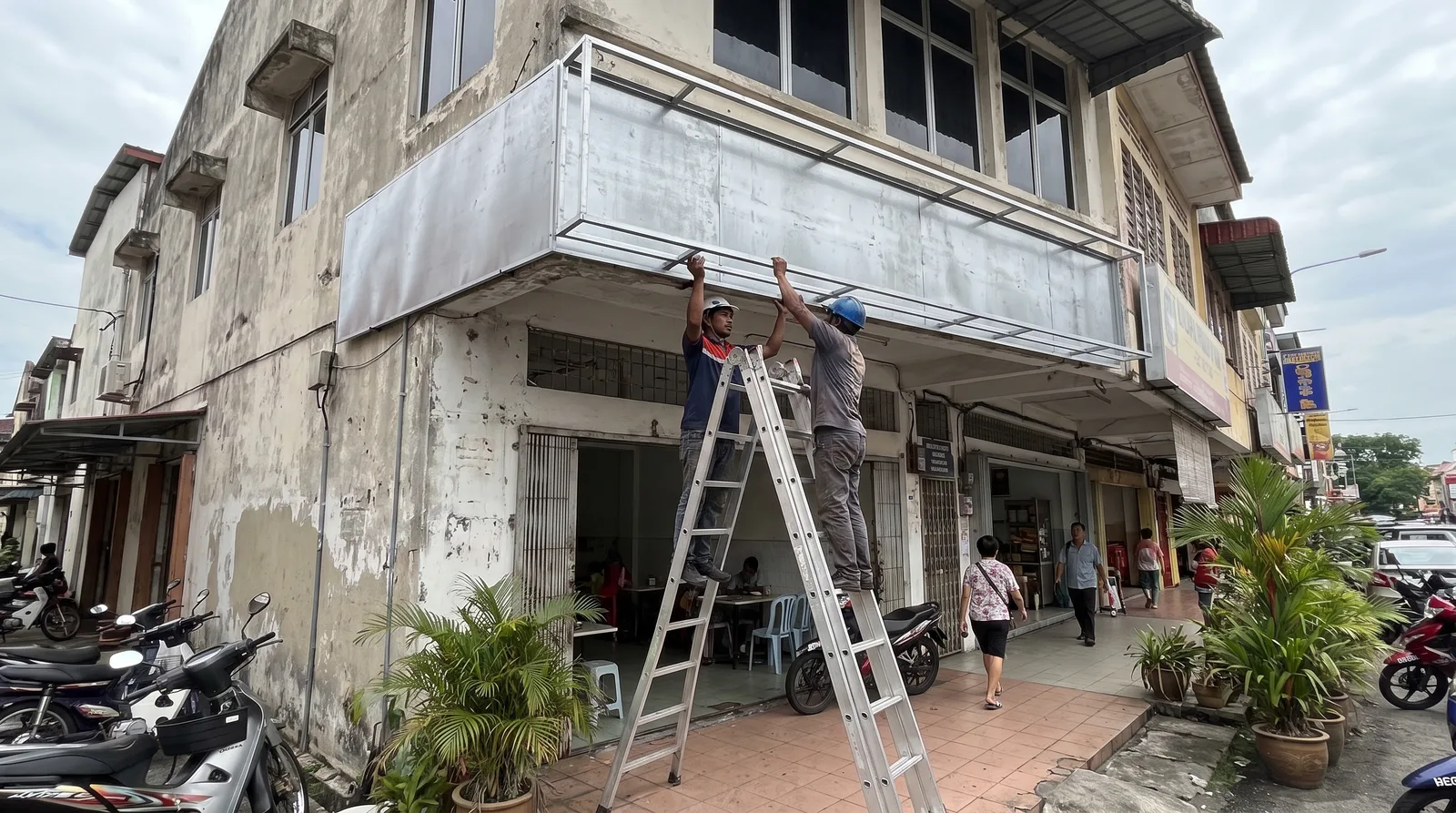 shopfront fascia signboard being installed by two workers on an aluminium ladder outside a Malaysian shoplot