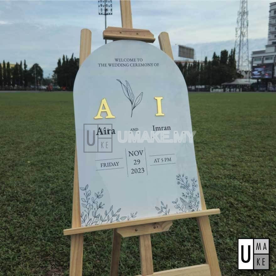 Arched acrylic wedding welcome board with gold mirror initials, standing on a wooden easel on a lawn